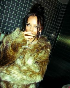 a woman in a fur coat holding a wine glass up to her mouth and looking at the camera