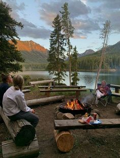 three people sitting around a campfire in the woods with mountains in the back ground