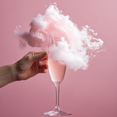 a hand holding a wine glass filled with pink liquid and white clouds floating out of it