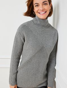 a woman wearing a gray turtle neck sweater and black pants, leaning against a white wall