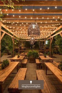 an outdoor dining area with wooden benches and lights hanging from the ceiling over it, surrounded by greenery