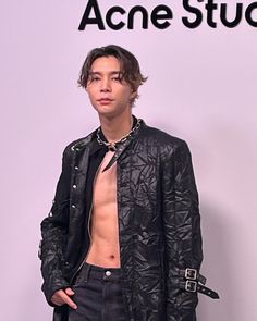 a man with no shirt standing in front of a wall wearing a black jacket and jeans