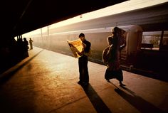 two people standing next to each other at a train station while the sun is setting