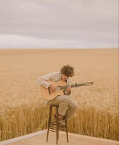 a man sitting on top of a wooden chair holding a guitar in front of a wheat field