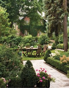 a table and chairs in the middle of a garden with trees, bushes and flowers