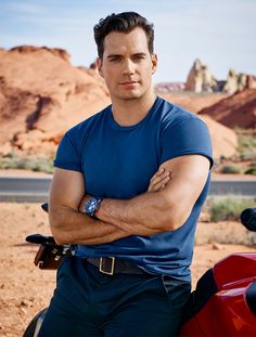 a man with his arms crossed standing next to a red motorcycle in front of mountains