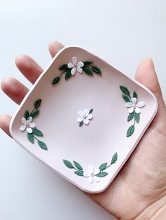 a hand holding a small white plate with green leaves and flowers painted on it's sides