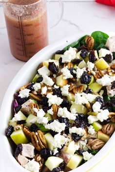 a salad in a white bowl next to a drink on a marble counter top with pecans, apples, and blueberries