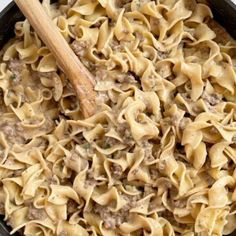 a skillet filled with pasta and meat on top of a white table next to a wooden spoon