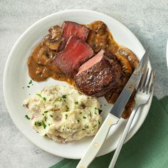 a white plate topped with meat, mashed potatoes and mushroom gravy next to a fork