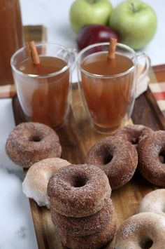 several donuts are on a wooden tray with cups of tea and apples in the background