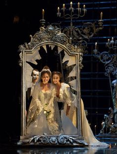 two women in white dresses standing on a stage with a large mirror and chandelier behind them