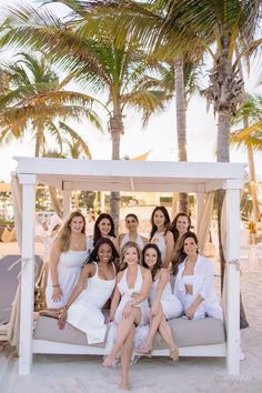 a group of women sitting on top of a white couch under a palm tree at the beach