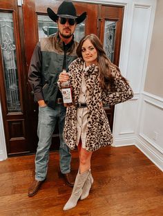 a man standing next to a woman in a leopard print coat and cowboy hat holding a bottle