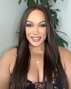 a woman with long brown hair wearing a black top and smiling at the camera while standing in front of a potted plant