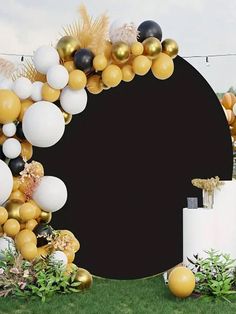 a black and gold balloon arch with white balloons on the top is surrounded by greenery