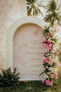 a wedding sign with flowers and greenery on the grass next to it in front of palm trees