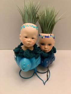 two small dolls sitting on top of a blue elephant head planter with grass growing out of it