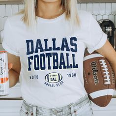 a woman holding a football and a can of soda in her right hand while wearing a white shirt with the word dallas football on it