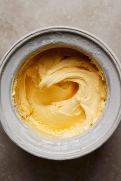 a metal bowl filled with yellow cream on top of a gray countertop next to a wooden spoon