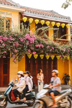 three people riding motorcycles in front of a yellow building with pink flowers on the balcony