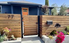 a wooden fence in front of a blue house with flowers on the ground and potted plants next to it