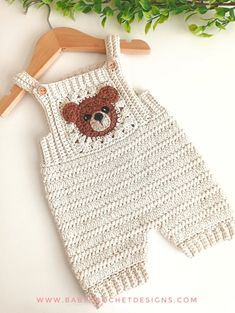 a crocheted bear overall is hanging on a wooden hanger next to some green leaves