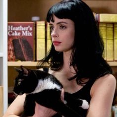 a woman holding a black and white cat in front of a book shelf with books on it