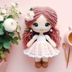 a crocheted doll sitting next to a cup of tea and flowers on a pink surface