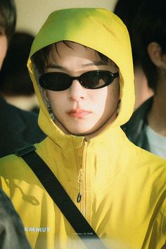 a person wearing sunglasses and a yellow raincoat is looking off to the side with other people in the background