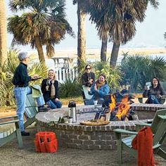 a group of people sitting around a fire pit in front of palm trees on the beach