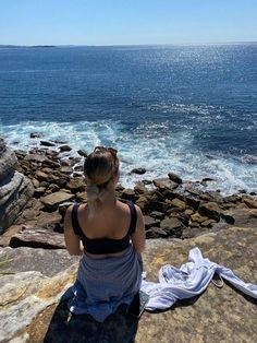 a woman sitting on top of a large rock next to the ocean and looking at the water