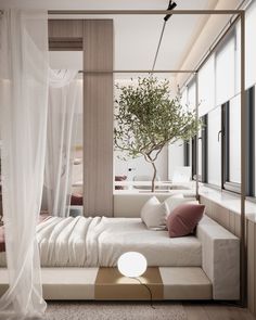 a white bed sitting under a window next to a tree in a room with lots of windows