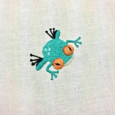 a close up of a small embroidered frog on a white fabric material with orange eyes