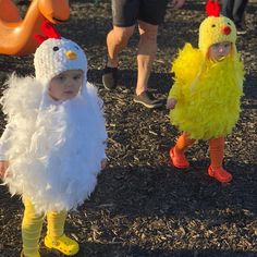 two small children dressed up as chickens and roosters at an outdoor play area with people in the background