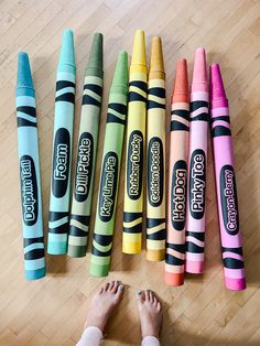 several crayons are lined up on the floor with one person's feet next to them