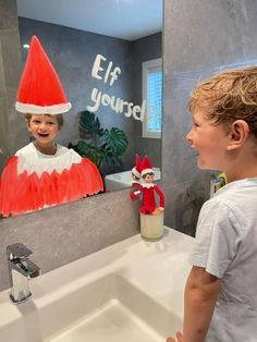 a young boy standing in front of a bathroom sink with an elf on the mirror