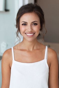 a smiling woman in a white tank top is looking at the camera and has her hair pulled back