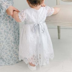 Handmade in the USA The Olivia dress features a delicate floral pattern lace and is lined with soft white linen. It has sheer lace sleeves that fall just above the elbow and is accented with a rose lace waist detail and a pale blue silk ribbon tie for a perfect fit. Trimmed with a petite floral edge lace, the dress falls between mid-calf to ankle length depending on height. It is finished with a timeless scalloped lace hem and a button closure for a secure and comfortable fit. To complete the lo Short Sleeve Delicate Lace Dress, Spring Lace Dress For First Communion, Spring Fitted Lace Dress For First Communion, Spring First Communion Fitted Lace Dress, Fitted Lace Dress For First Communion In Spring, Spring First Communion Lace Dress, First Communion Lace Dress With Lace Patchwork, White Lace Dress With Lace Bodice For Garden Party, Elegant Lace Trim Dress For Ceremony