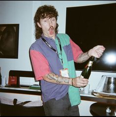 a man holding a bottle in his right hand and making a funny face while standing next to a table