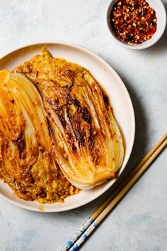 a white plate topped with cabbage covered in sauce next to chopsticks and a bowl of seasoning