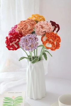 a white vase filled with colorful crocheted flowers next to a cup and saucer