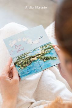 a woman is holding up a card with an image of the ocean and land on it