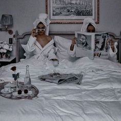 two women in bathrobes reading magazines on a bed with white sheets and pillows