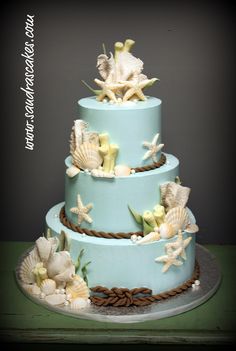 a three tiered blue cake with sea shells on the top and bottom, sitting on a green table