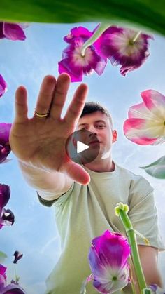 a man holding his hand up in the air with purple flowers around him and blue sky