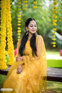 a woman in yellow sitting on a wooden bench with flowers hanging from the trees behind her