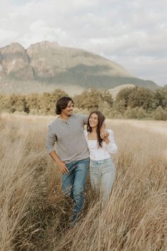 a man and woman standing in tall grass with mountains in the background