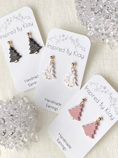 three christmas tree shaped earrings sitting on top of a white table next to some snowflakes