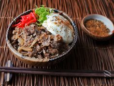 a bowl filled with meat and vegetables next to chopsticks on top of a mat